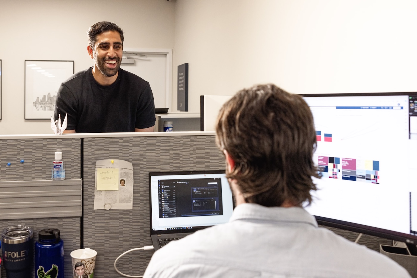 Two colleagues in an office cubicle; one working at his computer, another smiling in the background