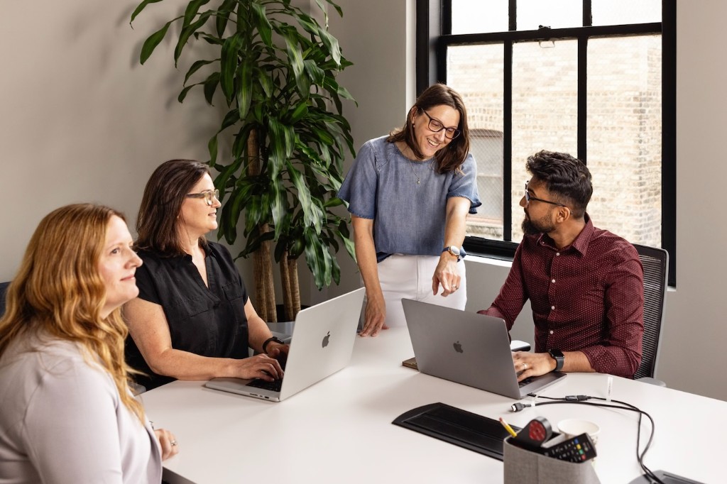 Four colleagues collaborating around a table with laptops in a bright office